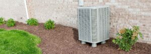 Air conditioner sitting in a garden with woodchips and green plants next to a brick building
